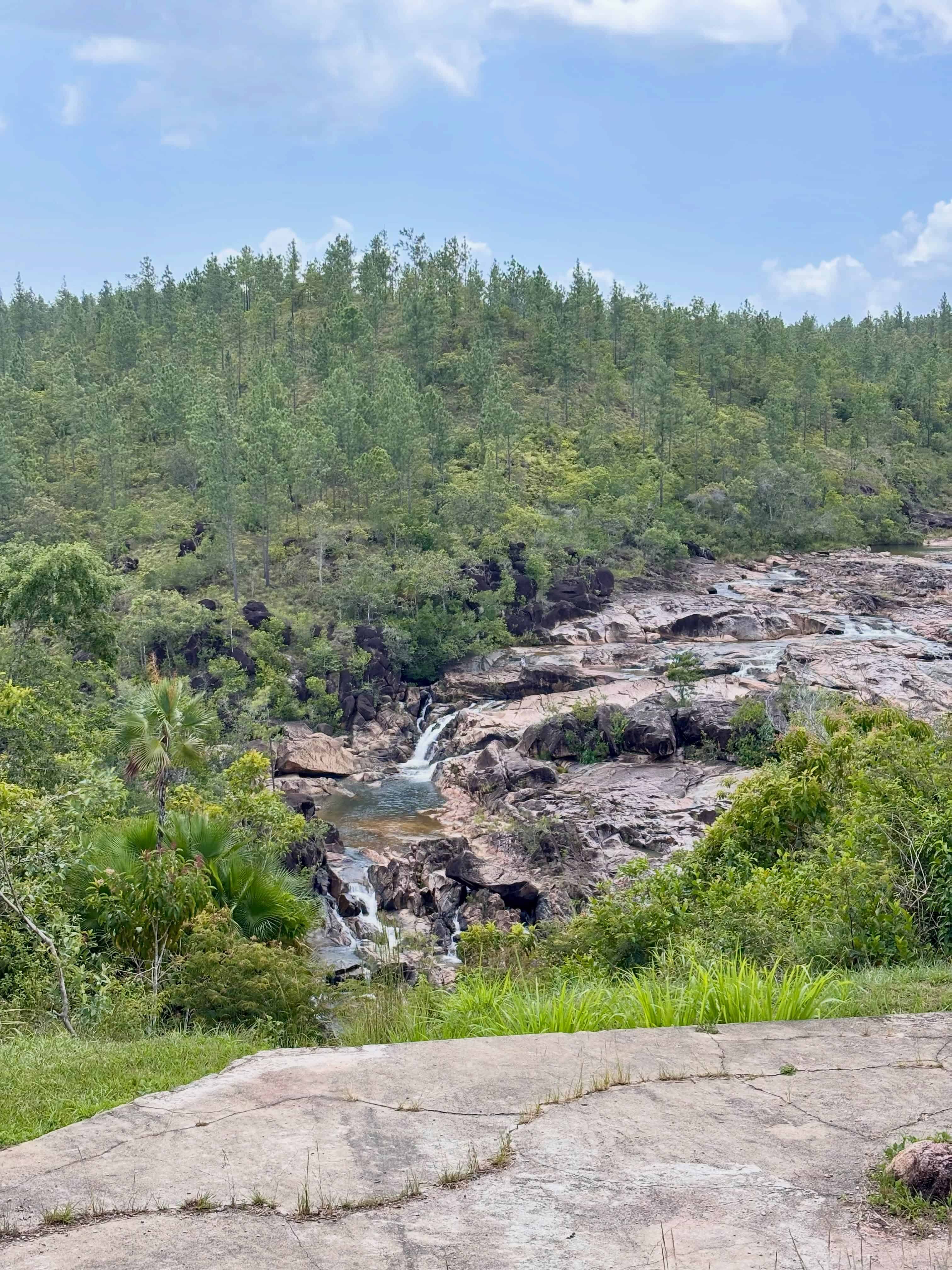 Water flows over rocky terrain, surrounded by green forest under a blue sky with light clouds&mdash;an idyllic scene reminiscent of a Belize reef and ruins trip.