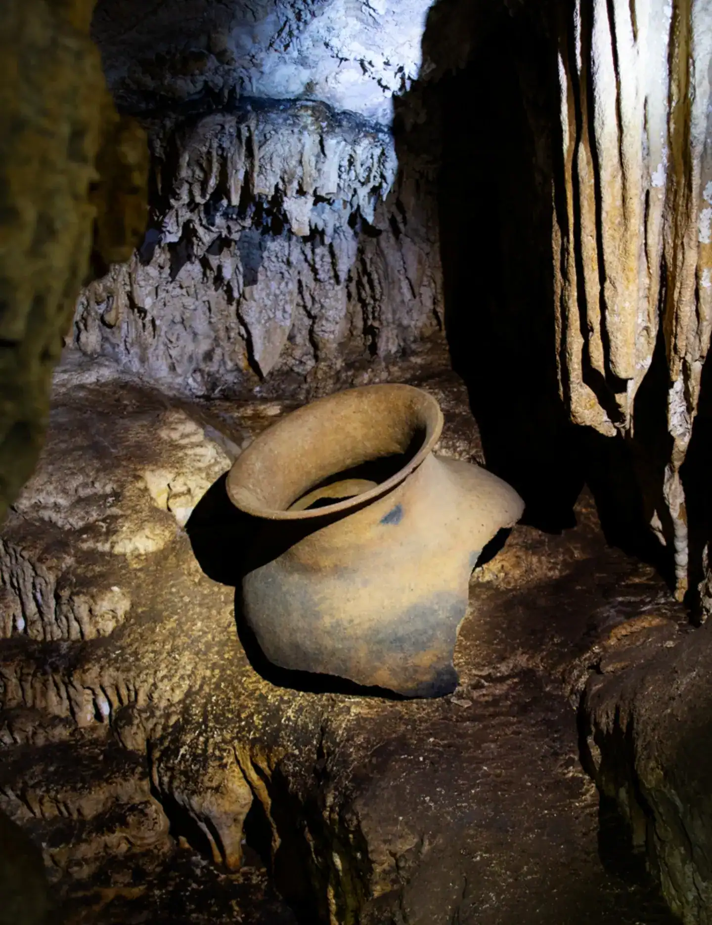 An old, broken clay pot lies on the rocky floor of a dimly lit cave with stalactites and rough walls, reminiscent of artifacts discovered during a Belize reef and ruins trip.