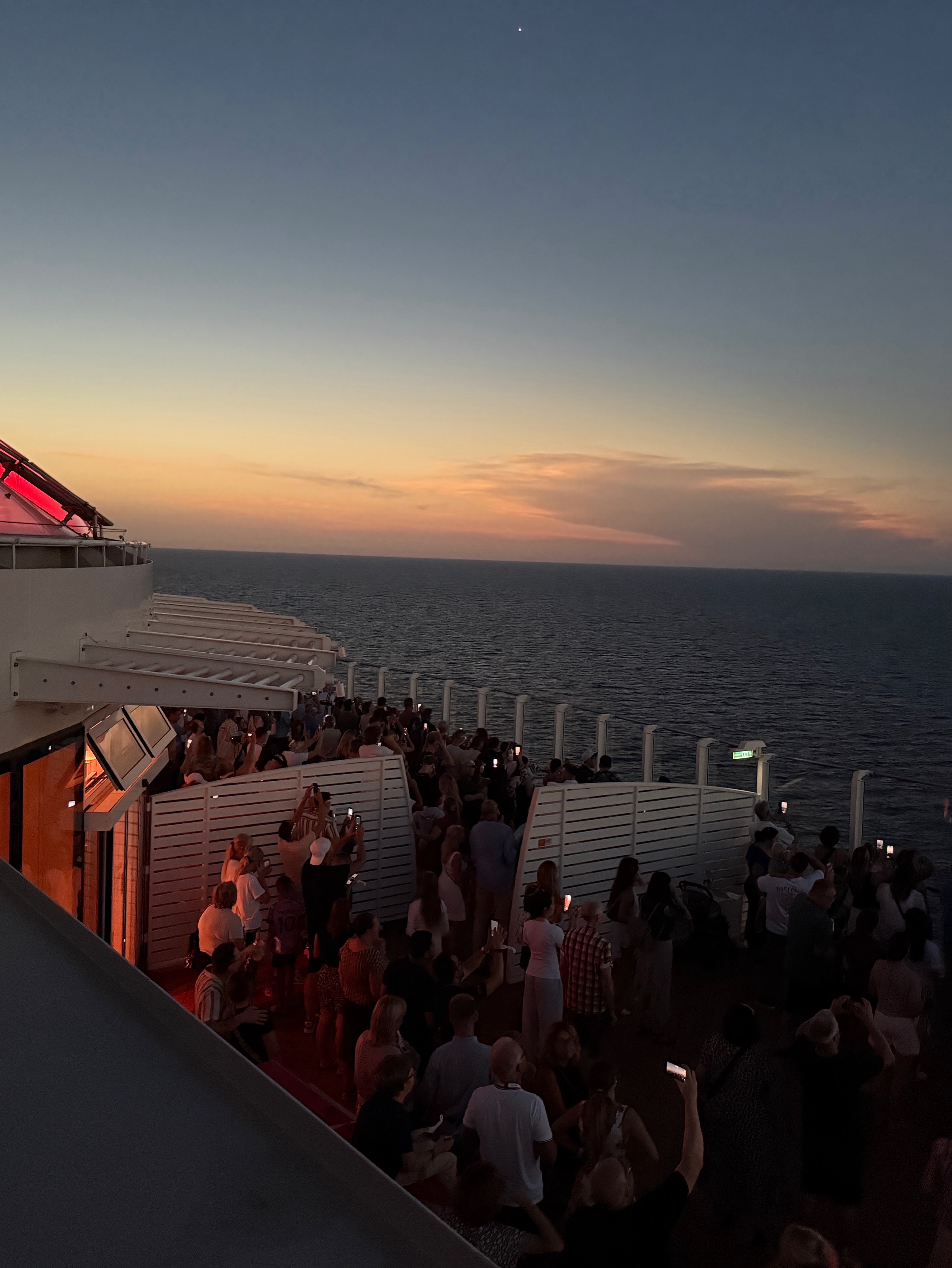 Crowd gathers on a ship deck at sunset during a Solar Eclipse Cruise, many people taking photos over the ocean.