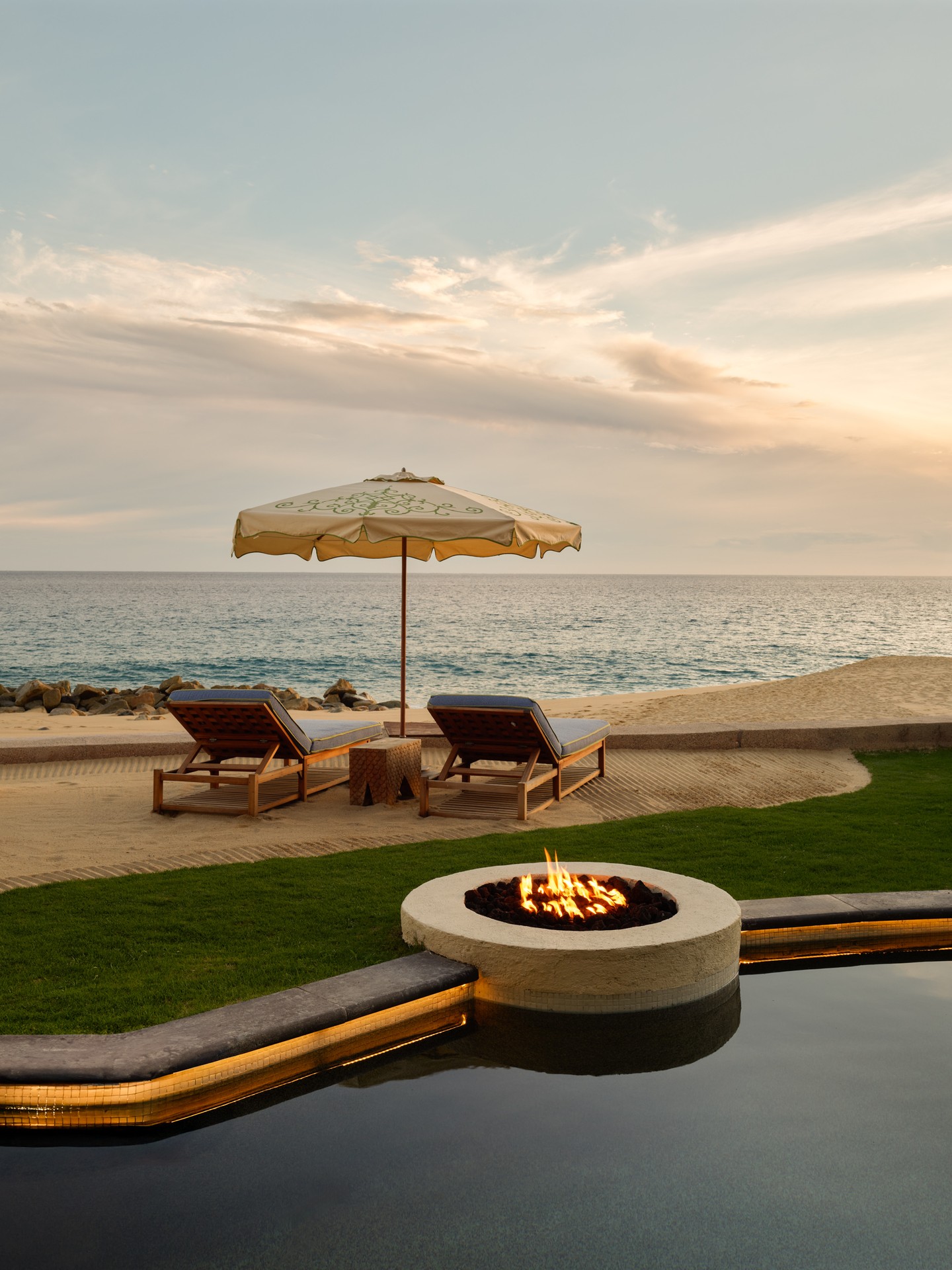 Two lounge chairs and an umbrella face the ocean near a fire pit and a pool at sunset, evoking the relaxed charm of sought-after European destination dupes.