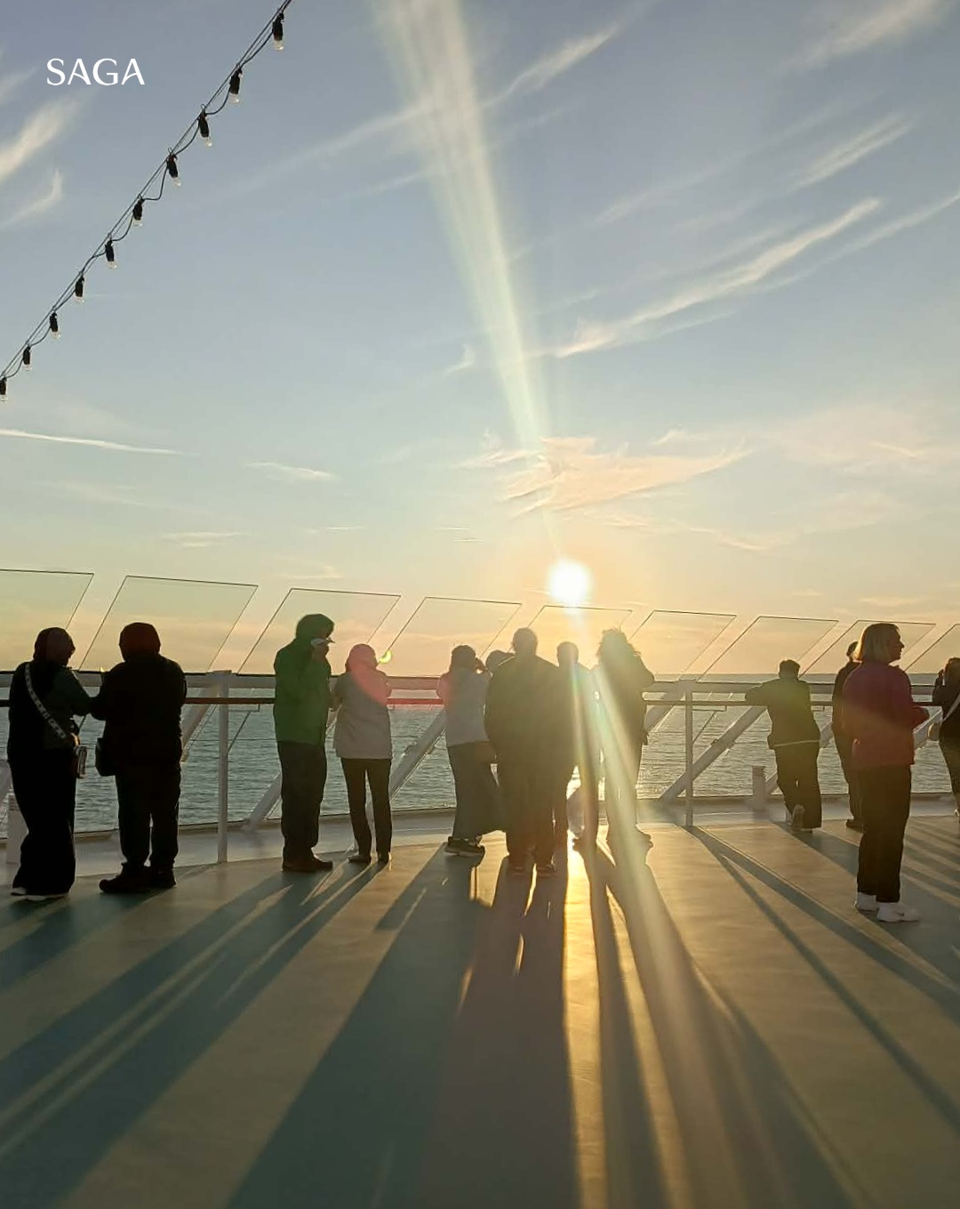 People stand on a ship deck at sunset, with long shadows and sunlight reflecting on the floor, enjoying the mesmerizing views during a Solar Eclipse Cruise.