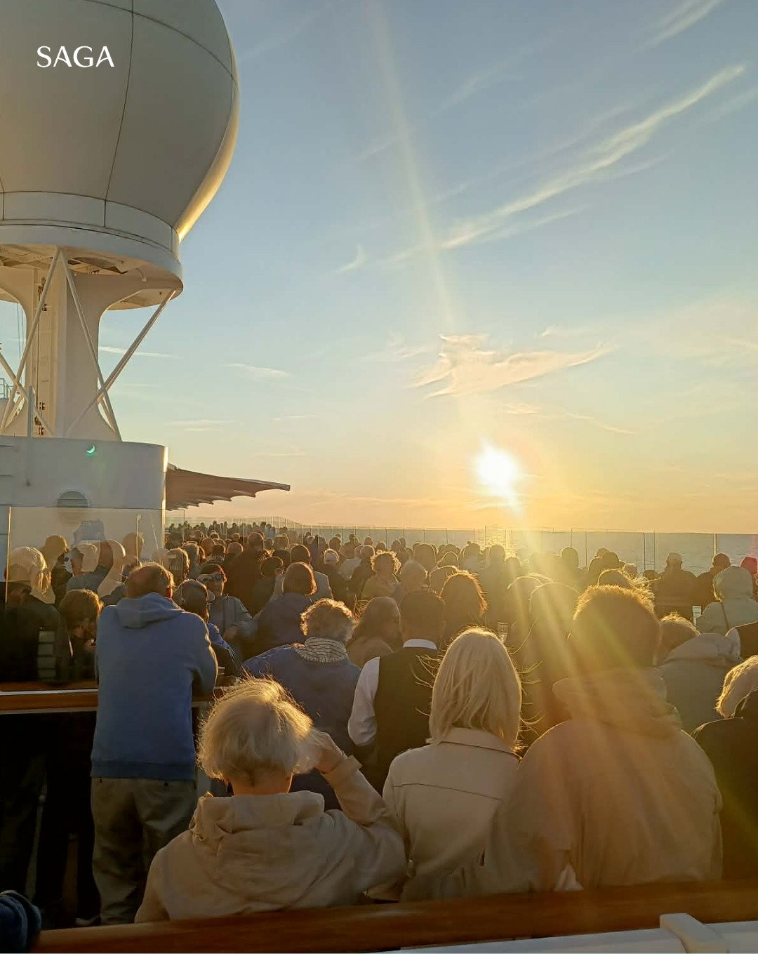 Crowd of people on a ship deck at sunset, sunlight streaming, with &ldquo;SAGA&rdquo; text in the sky&mdash;capturing the magical atmosphere of a Solar Eclipse Cruise.