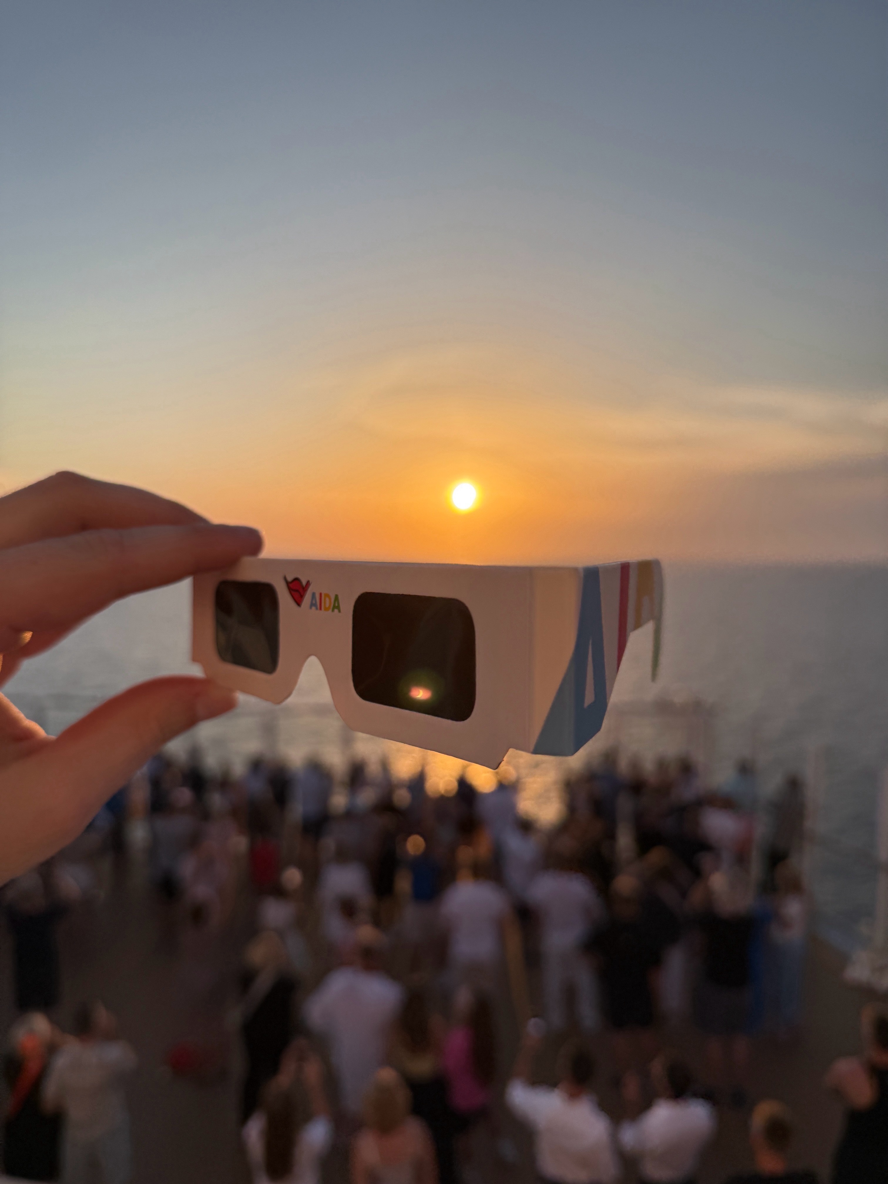 A hand holds 3D glasses up to a sunset over the ocean, with a crowd gathered on a deck below during a Solar Eclipse Cruise.