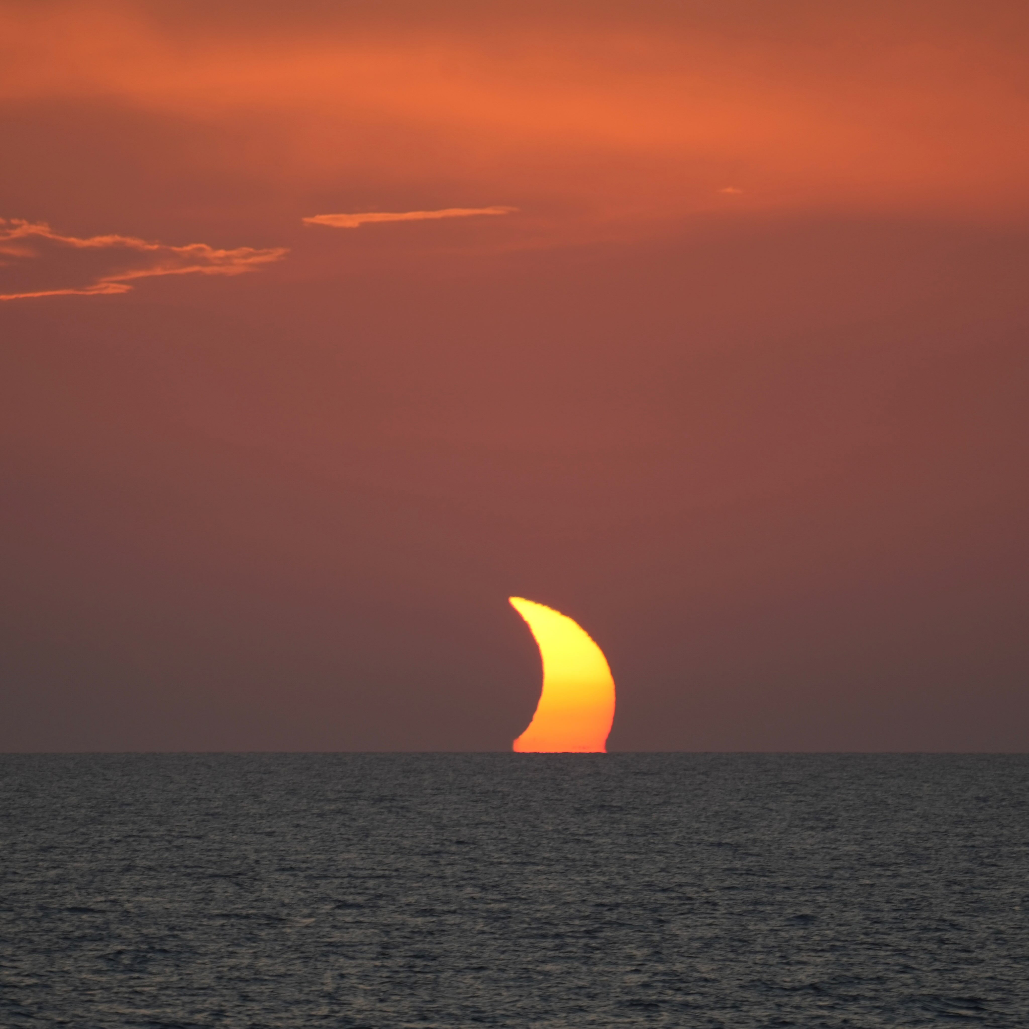 Partial sun sets over calm ocean, casting orange hues across the sky and reflecting light on the water&mdash;a tranquil scene reminiscent of a Solar Eclipse Cruise experience.