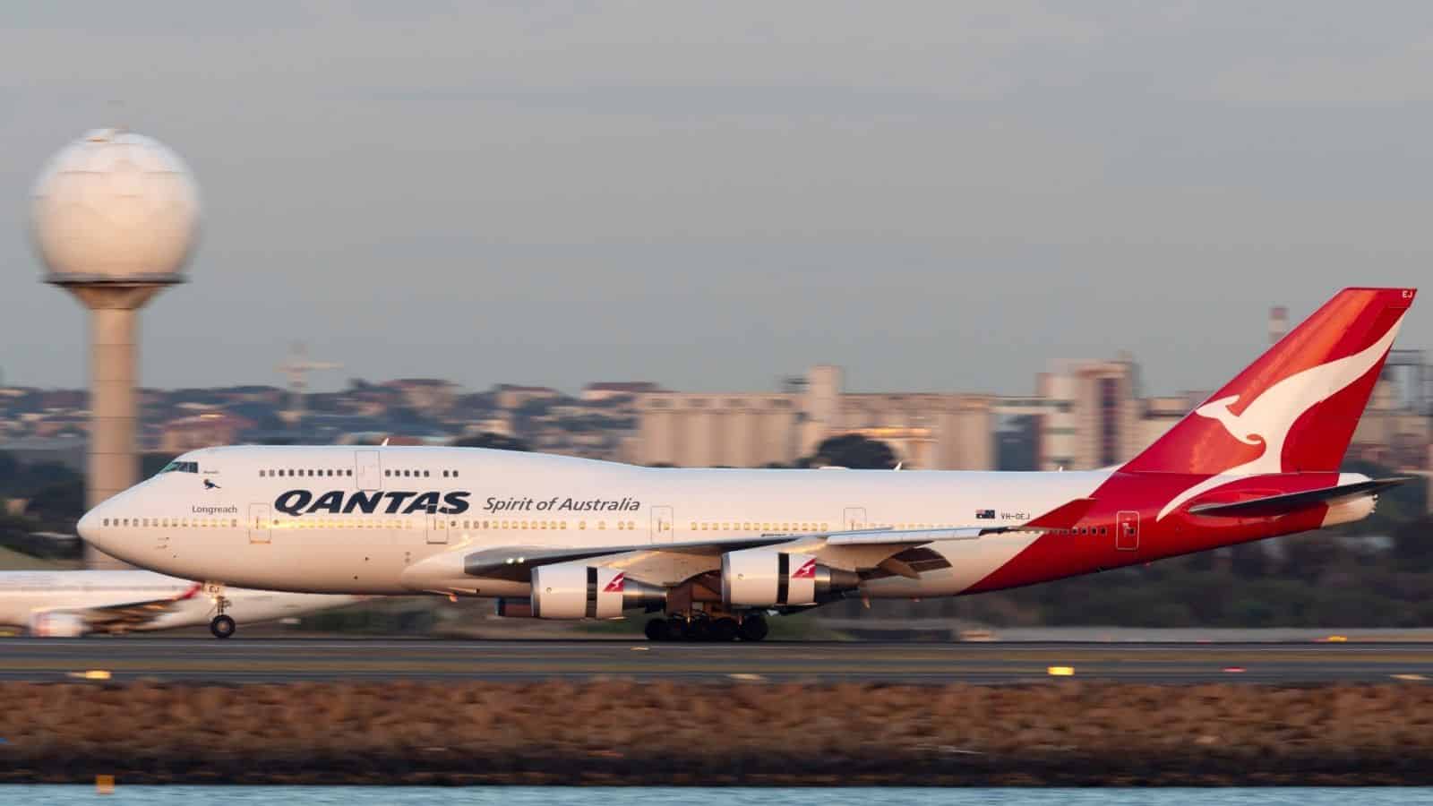Qantas Boeing 747 airplane landing on a runway with a cityscape and radar tower in the background.