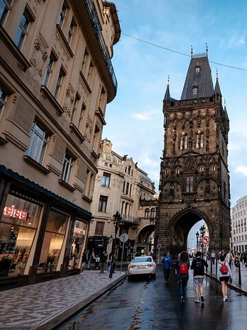 People walk on a street leading to a tall, historic tower in a European city with cloudy skies above.