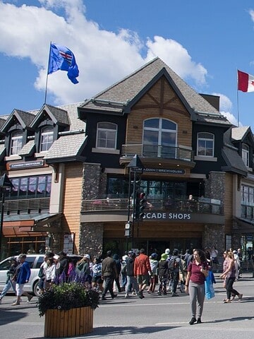 People cross a street in a mountain town with shops; a Canadian flag waves atop a building.