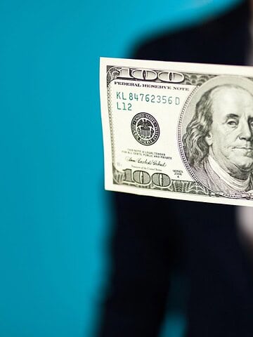 Person holding a $100 bill in front of a blue background, focus on the money.