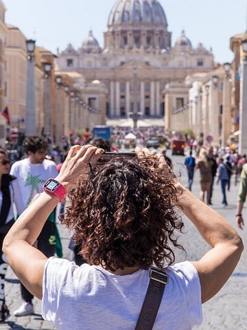 Person with curly hair faces busy street toward St. Peter’s Basilica in Vatican City on a sunny day.