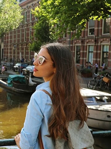 Woman with long hair stands on a bridge overlooking a canal lined with boats and trees in a city.