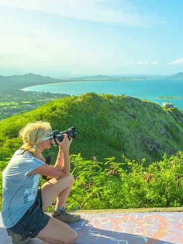 A person takes a photo on a hilltop overlooking a coastal town and turquoise ocean under a clear sky.