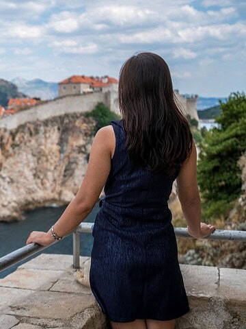 Woman in navy dress overlooks a historic coastal city with stone walls and red rooftops under a blue sky.