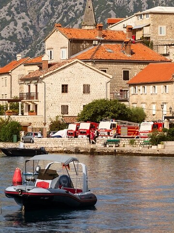 A small boat floats on water near stone buildings with red roofs and green hills in the background.