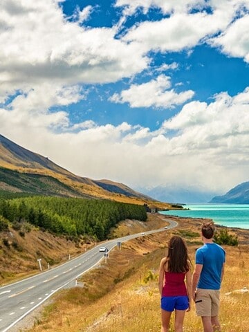 A couple stands by a winding road, overlooking a turquoise lake and mountains under a partly cloudy sky—capturing the natural beauty often found in easy travel destinations.