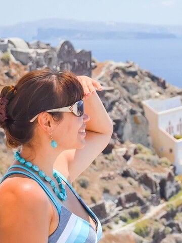 Woman in sunglasses and blue necklace looks out over a sunny seaside cliff with white buildings and blue water.