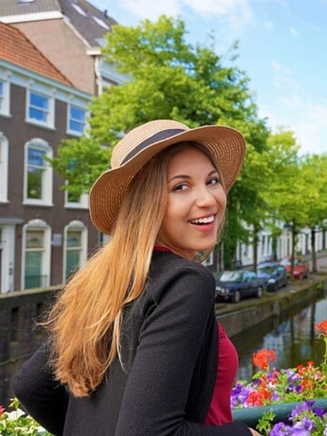Smiling woman in a hat stands by colorful flowers on a canal with trees and buildings in the background.