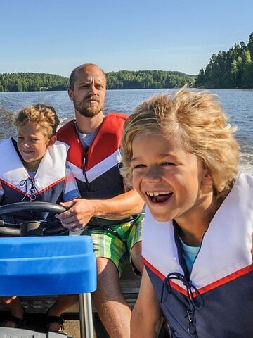 A man and two smiling children in life jackets ride a motorboat on a sunny lake.