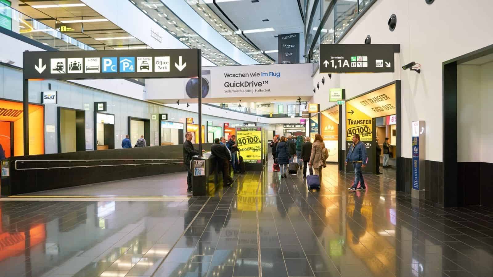 People walk through a modern airport terminal with signs, shops, and bright lights visible.