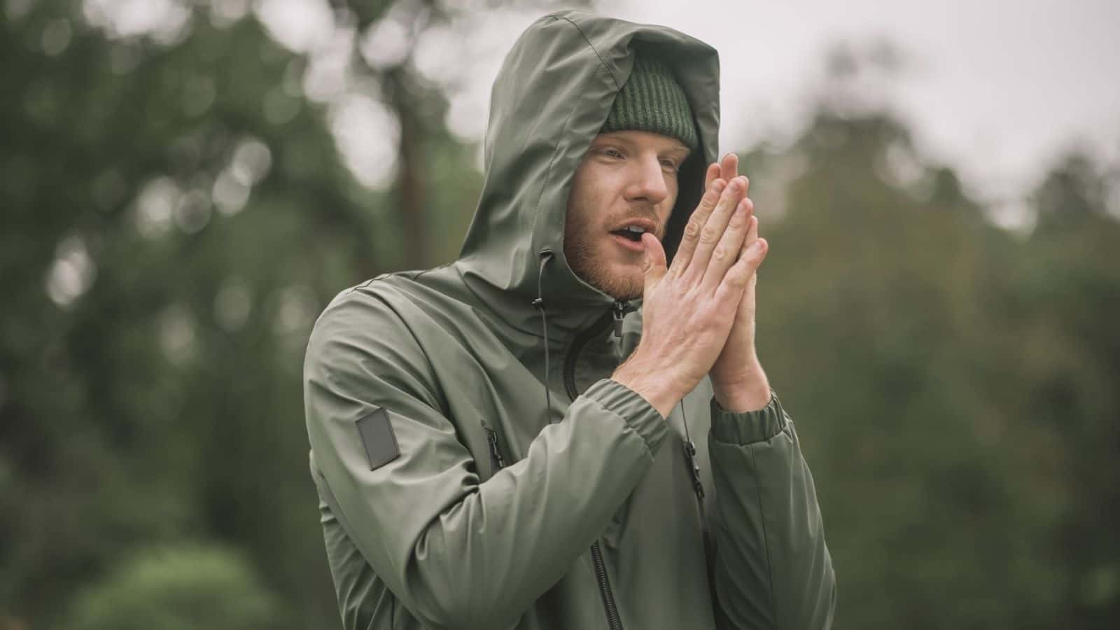 Man in a green hooded jacket and beanie stands outdoors, warming his hands near his face—ready for hiking in unpredictable weather with all his travel essentials close by.