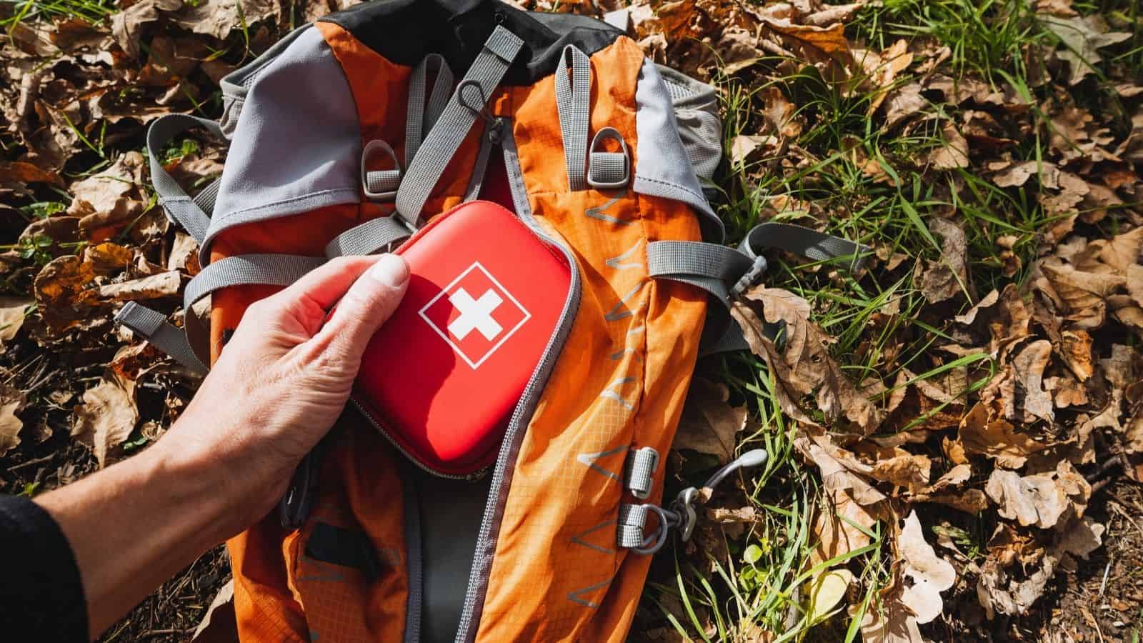 Hand placing a red first aid kit—one of the travel essentials for hiking—on an orange backpack lying on leaves and grass outdoors, ready to tackle unpredictable weather.