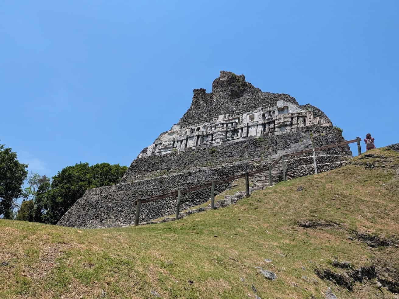Ancient stone Mayan temple with carvings stands on a grassy hill under a clear blue sky&mdash;an unforgettable highlight of any Belize reef and ruins trip.
