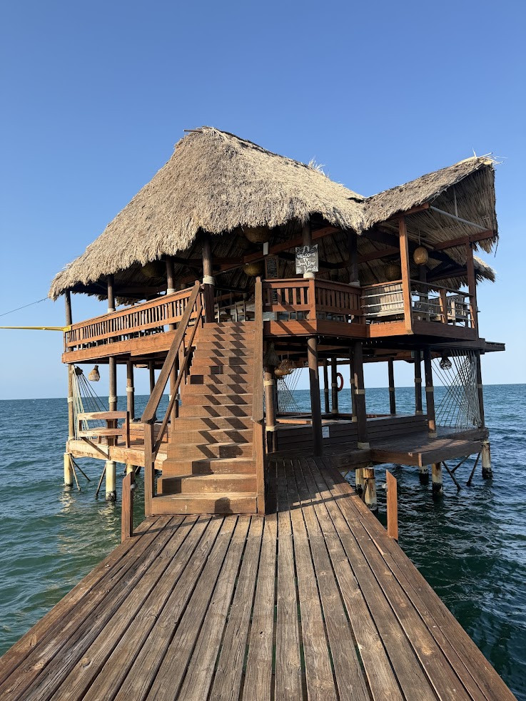 Wooden hut with a thatched roof on stilts over ocean water, viewed from a dock under a clear blue sky&mdash;a scene reminiscent of the tranquil escapes often mentioned in the Lodge at Jaguar Reef review.