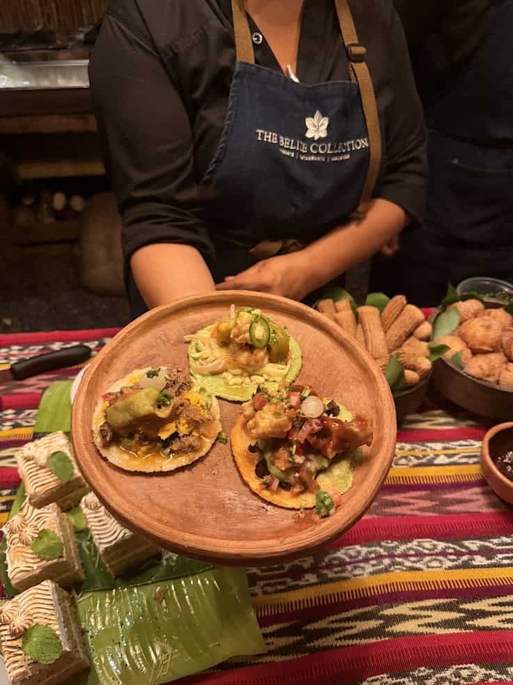 A person holds a wooden plate with three assorted tostadas; desserts are on a colorful tablecloth below, reminiscent of the vibrant dining experiences mentioned in The Rainforest Lodge at Sleeping Giant Review.