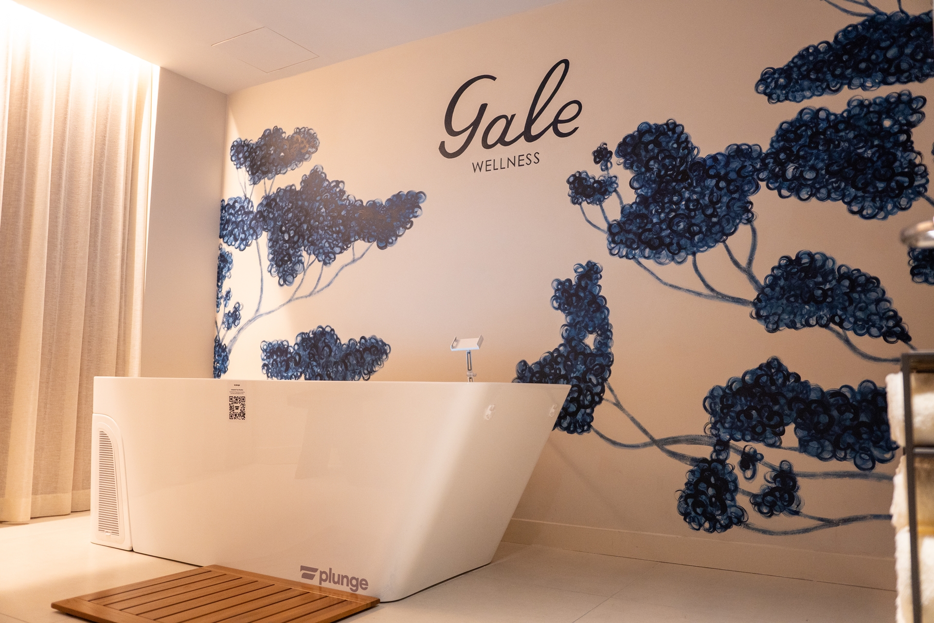 Modern white soaking tub by a wall with “Gale Wellness” and blue floral mural, light streaming from left—an inviting scene reflecting the serene atmosphere featured in many Gale Miami Hotel & Residences Review highlights.