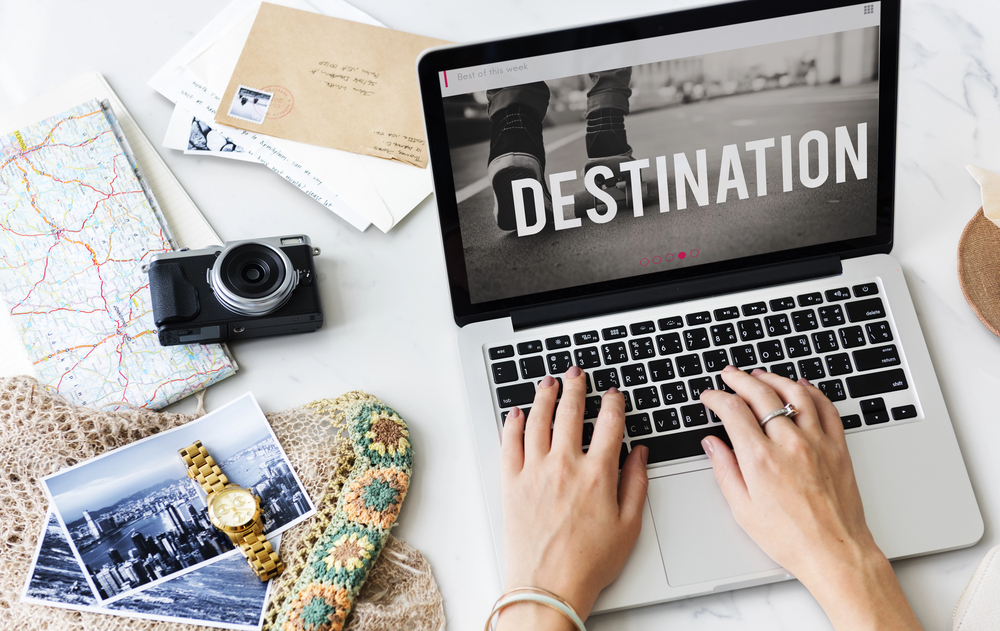 Hands on laptop with “DESTINATION” on screen, surrounded by travel items, photos, a map, and hints of planning to travel with teens.