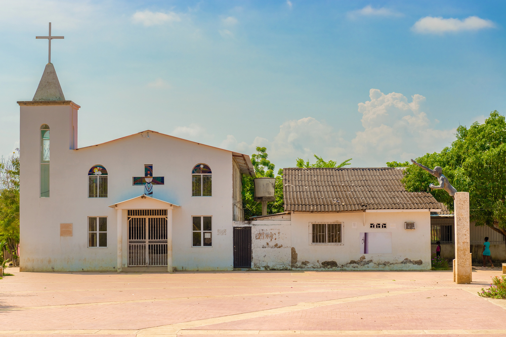 A small white church and nearby building under a blue sky with trees and a statue in the foreground, reminiscent of the charming historic sites you might discover while exploring things to do in Cartagena.