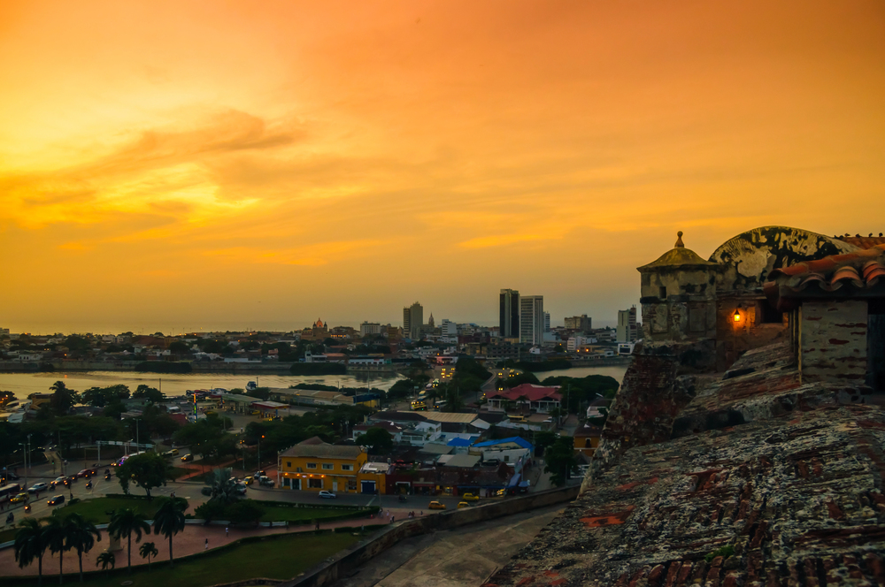Sunset over a cityscape with a historic stone fort, colorful buildings, and a distant modern skyline&mdash;an iconic view that captures the charm of Cartagena. Among the many things to do in Cartagena, watching this breathtaking sunset from the ancient fort walls is an experience not to be missed.