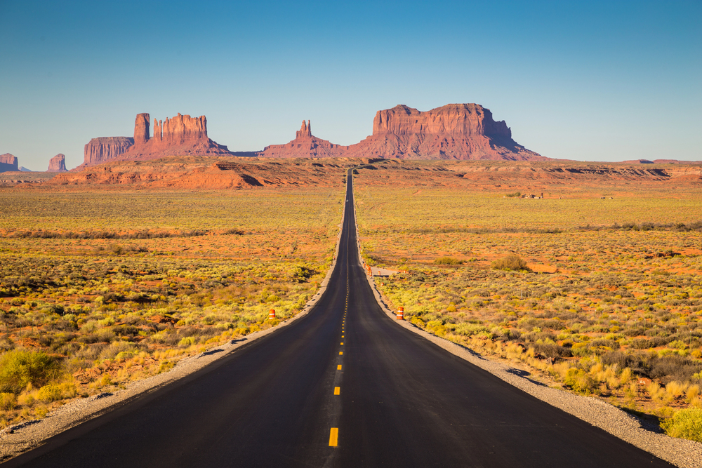 A straight road leads through desert toward distant red rock mesas under a clear blue sky, making it the perfect route to travel with teens and create unforgettable family memories.