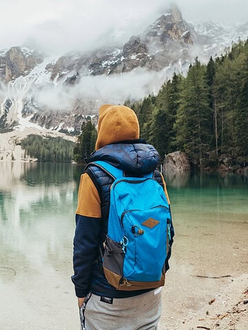 A person with a blue backpack stands by a clear mountain lake, surrounded by trees and snowy peaks, embracing the spirit of empty nest travel.