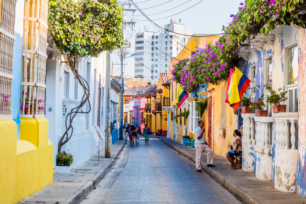 Colorful street with flowers, people, and Colombian flags, with buildings lining both sides of the road&mdash;a perfect scene that showcases some of the vibrant things to do in Cartagena.