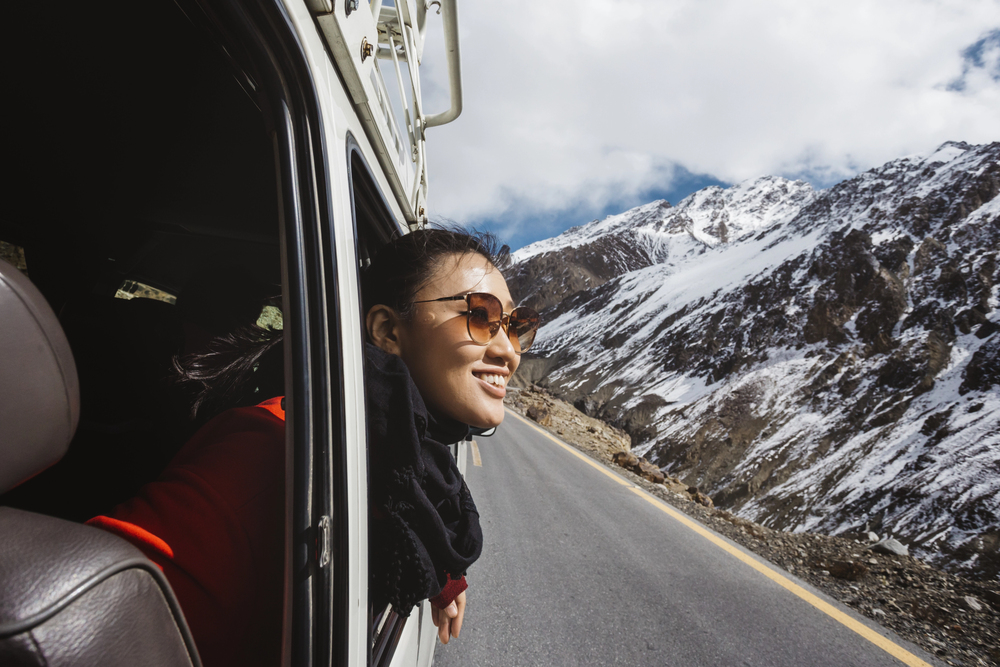 Woman in sunglasses leans out car window, smiling, with snowy mountains and blue sky in the background&mdash;embracing the freedom of empty nest travel.