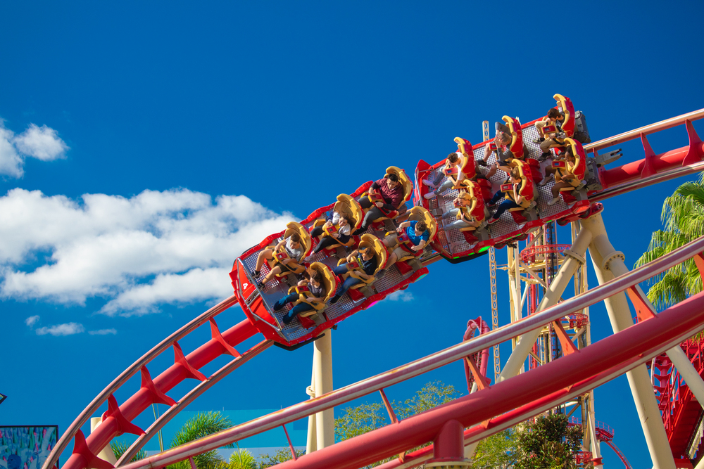 People ride a red roller coaster under a bright blue sky with a few white clouds and green trees nearby—a thrilling highlight among the top things to do in Orlando with kids.