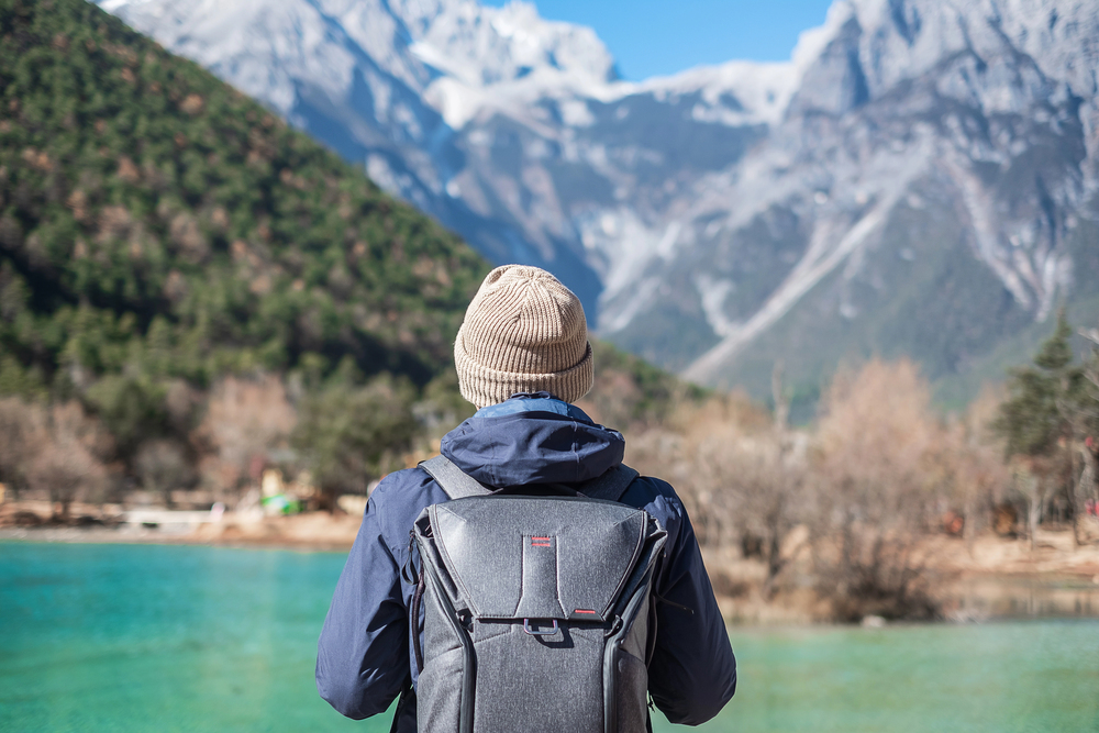 A person with a backpack faces a turquoise lake and snowy mountains under a clear blue sky, embracing the spirit of empty nest travel and new adventures.
