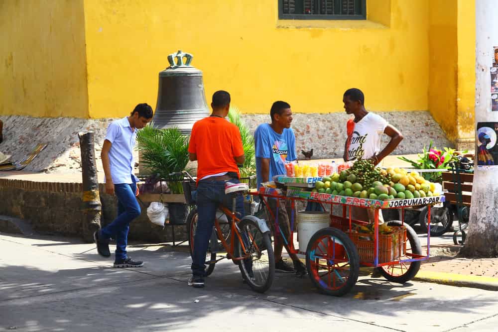 Four men stand by a colorful fruit cart on a sunny street near a yellow wall and a large bell&mdash;a lively scene that showcases one of the many authentic things to do in Cartagena.