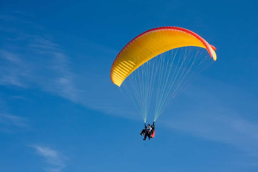 Two people paragliding together under a yellow and red parachute against a clear blue sky—an unforgettable adventure and one of the most exciting things to do in Medellin with kids.