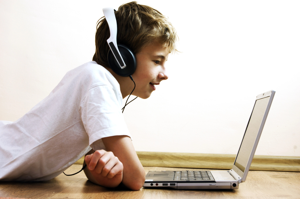 Boy wearing headphones lying on the floor, smiling and looking at a laptop screen in front of him—perfect for planning your next travel with teens adventure.