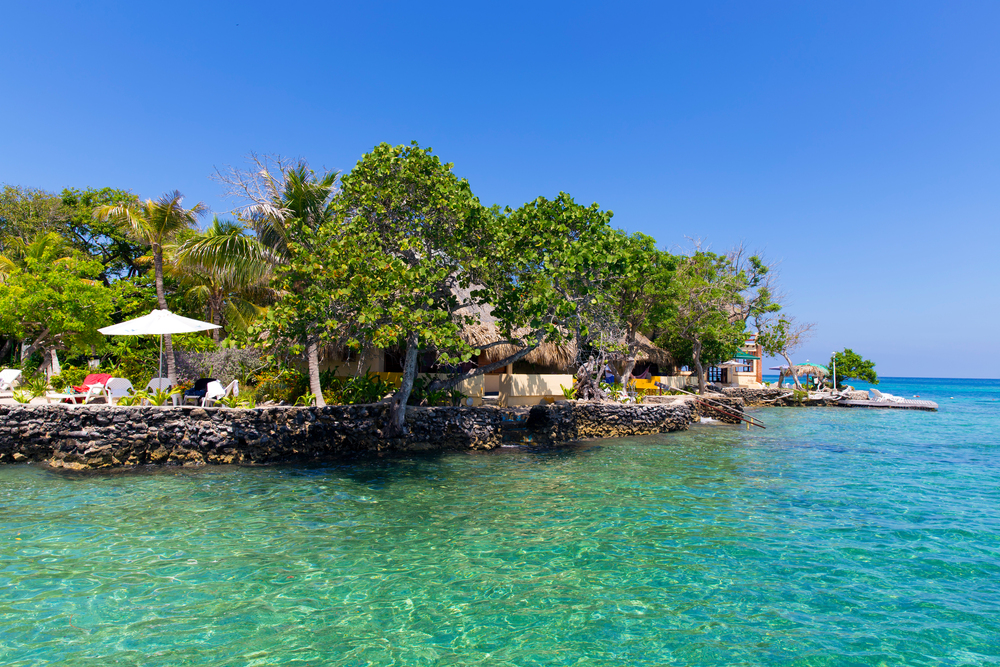 Tropical island with clear blue water, lush trees, and thatched-roof huts under a bright blue sky&mdash;perfect for relaxing or exploring unforgettable things to do in Cartagena.