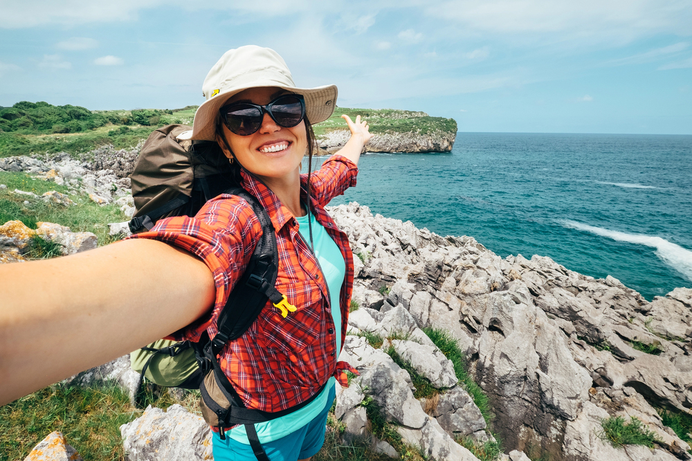 Smiling hiker in sunglasses poses on rocky coastline with blue ocean and cliffs in the background, embracing the spirit of empty nest travel.