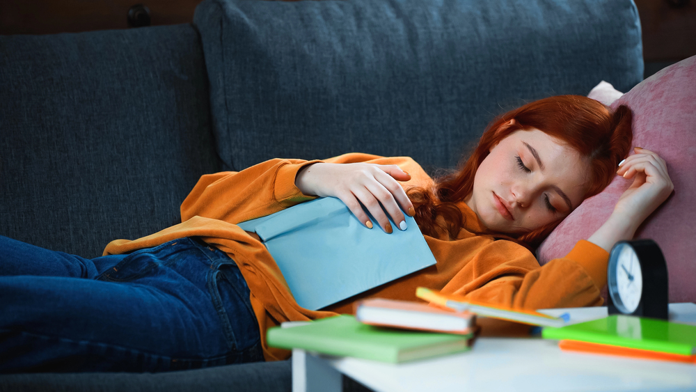 A girl with red hair naps on a couch, holding a book, with notebooks and a clock on the table nearby—perhaps dreaming of her next adventure as she plans to travel with teens.