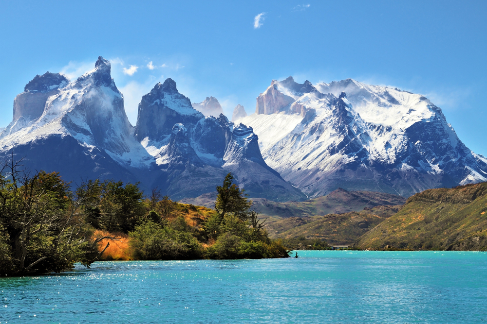 Snowy jagged mountains rise above turquoise water and green trees under a clear blue sky, creating an inspiring scene perfect for empty nest travel adventures.