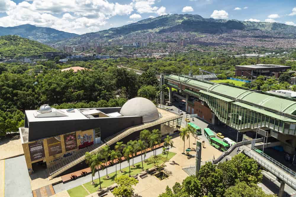 Aerial view of a metro station, dome building, and cityscape with green hills in the background—an inspiring glimpse of the vibrant landscape and urban attractions among the many Things to Do in Medellin with Kids.
