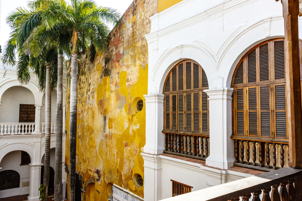 Tall palm trees beside a weathered yellow wall and white colonial-style arched windows and balconies create a picturesque scene&mdash;one of the many things to do in Cartagena for those who love exploring beautiful, historic architecture.