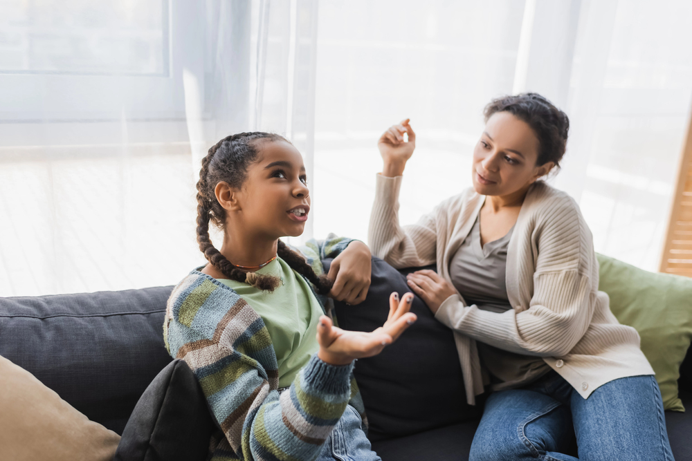A girl with braids talks about her experiences of travel with teens while a woman listens attentively on a couch in a sunlit room.