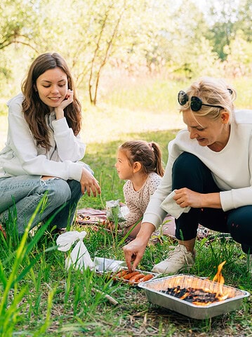 Three people cook food on a portable grill outdoors, smiling and enjoying a picnic in a grassy area&mdash;making the most of travel with teens and creating unforgettable memories together.