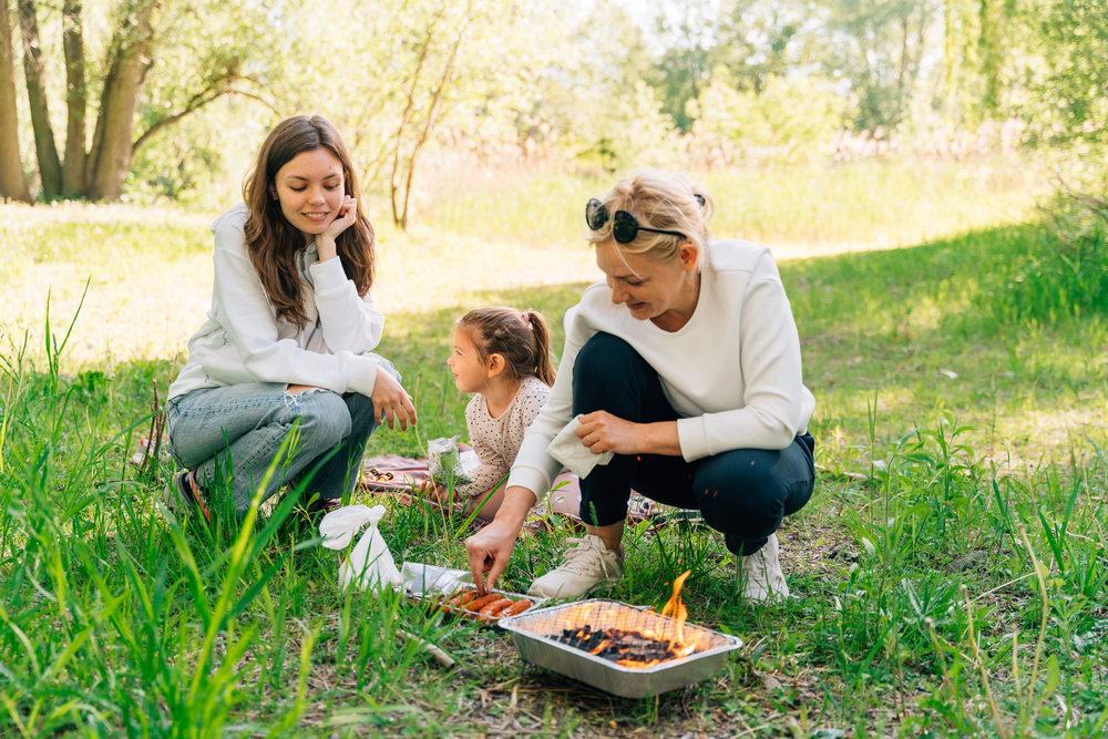 Three people cook food on a portable grill outdoors, smiling and enjoying a picnic in a grassy area—making the most of travel with teens and creating unforgettable memories together.