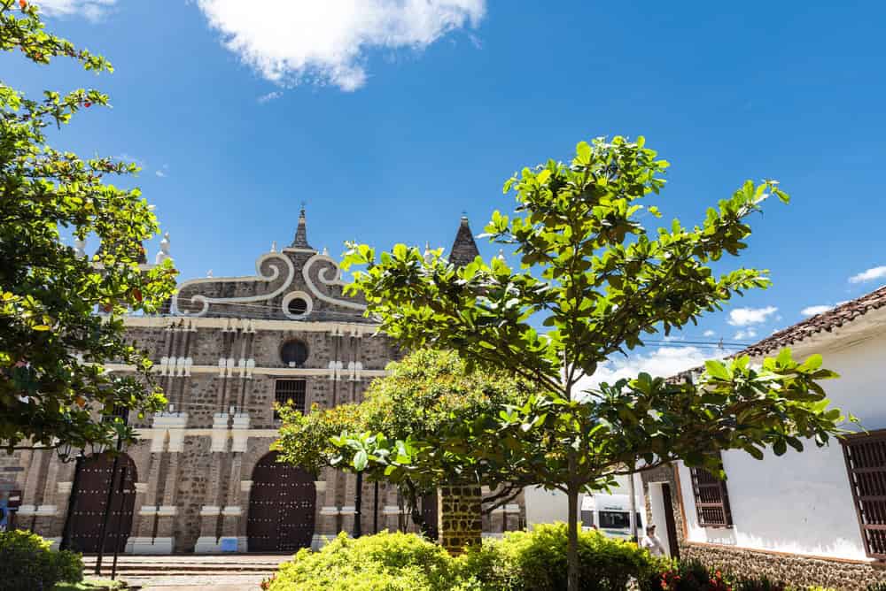 Colonial stone church with ornate facade, green trees, and a bright blue sky with clouds—a perfect spot to explore while discovering Things to Do in Medellin with Kids.